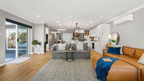 A modern living room with a grey sofa, a brown leather sofa, and a small round table. There is a kitchen area in the background with white cabinets and a stainless steel refrigerator.