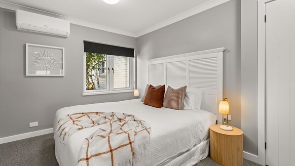 Une chambre à coucher avec un lit, une table de chevet avec une lampe, une fenêtre avec des stores et un climatiseur fixé au mur.