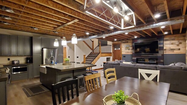 Eine moderne Küche mit einer zentralen Kochinsel, ein Essbereich mit Tisch und Stühlen, ein Wohnzimmer mit Sofa und Fernseher sowie eine Treppe zu einer oberen Ebene.
