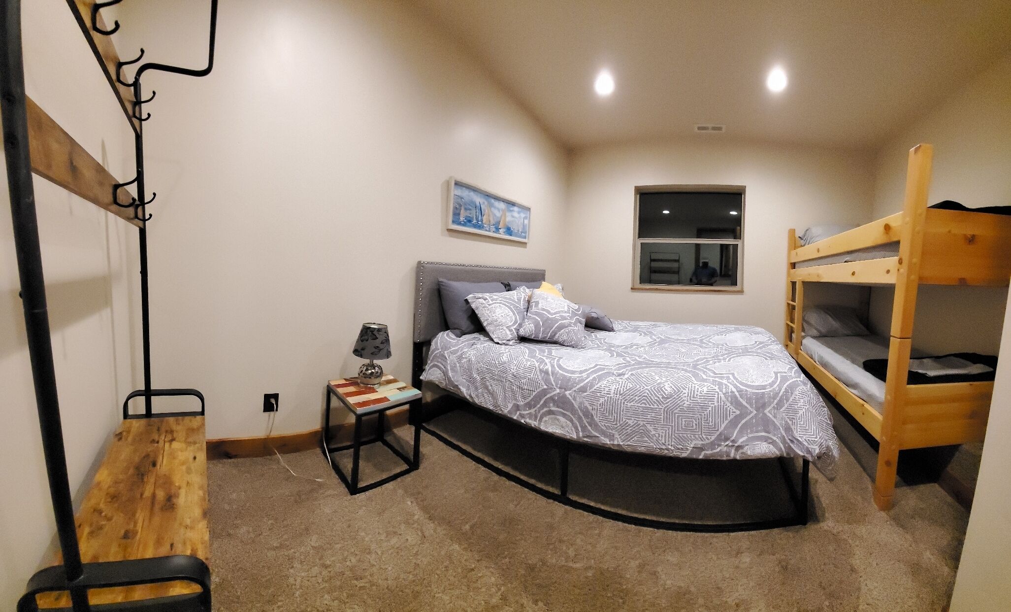 A bedroom with a bed, a bedside table with a lamp, a bunk bed, a window, and a painting on the wall.