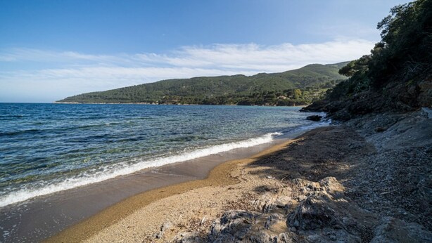 Una playa con agua cristalina, costa rocosa y colinas al fondo.