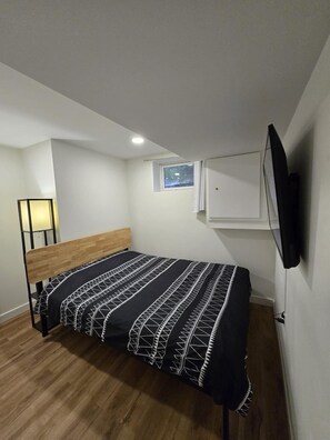 A small, modern bedroom with a single bed, a nightstand, a wall-mounted TV, and a small window. - New Modern 2 Bed Suite - Steps From West Edm Mall (Edmonton)