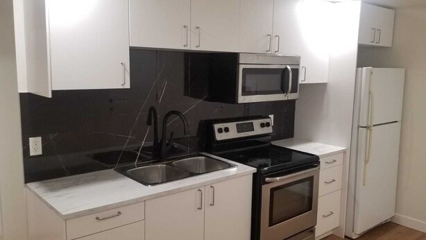 Cozinha compacta com armários brancos, bancada preta, pia de aço inoxidável, micro-ondas, forno e geladeira.