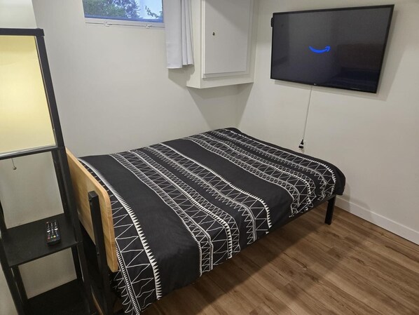 A small, neatly arranged bedroom with a single bed, a TV mounted on the wall, and a window with curtains. - New Modern 2 Bed Suite - Steps From West Edm Mall (Edmonton)