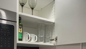 Uma cozinha com micro-ondas, chaleira e uma prateleira com copos de vinho e uma garrafa de azeite.