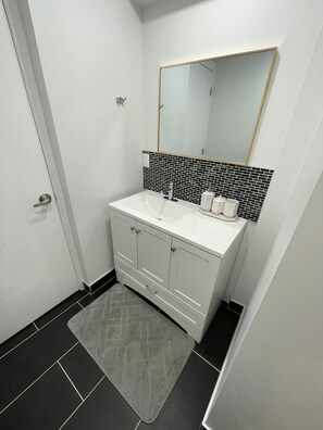 A bathroom with a white vanity, black and white tiled backsplash, and a gray rug. - New Modern 2 Bed Suite - Steps From West Edm Mall (Edmonton)