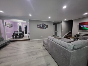 Un salon moderne avec un canapé gris, un coin repas agrémenté d’un lustre et un escalier menant à un autre niveau.