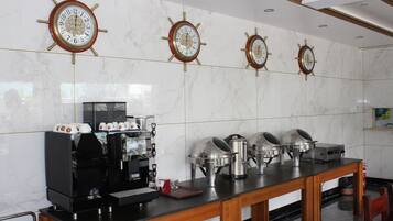 Una cocina de hotel con paredes de mármol, cuatro relojes de barco y equipo para servicio de buffet, además de una máquina de café.
