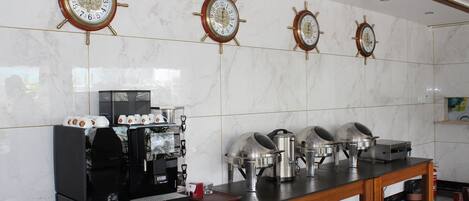 A hotel kitchen with marble walls, four ship's wheel clocks, a coffee machine, and buffet service equipment.
