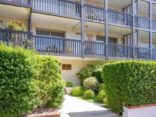 A multi-story residential building with balconies, a well-maintained garden, and a sign indicating the address.