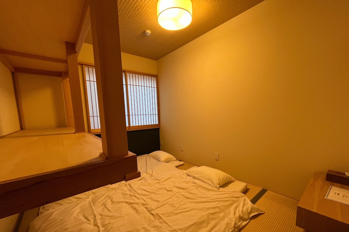 Habitación de hotel con literas, un escritorio con silla y una ventana con persianas verticales.