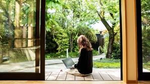 Eine Person sitzt auf einer Holzdielenveranda mit einem Laptop und blickt auf einen Garten mit Bäumen und einem Weg.