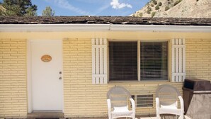 Un edificio de ladrillo amarillo con una puerta blanca, dos sillas de mimbre y una ventana con contraventanas.