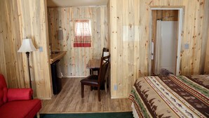 Une pièce aux murs lambrissés, un lit avec une literie à motifs, une chaise rouge, un bureau avec une lampe et un espace douche.