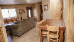 A living room with a sofa, a dining table with chairs, a window with blinds, and a door leading to another room. - Ponderosa Cabin (Deluxe) (Sevier)