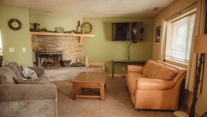 A living room with a stone fireplace, a television mounted on the wall, and a window with blinds. - Ponderosa Cabin (Deluxe) (Sevier)