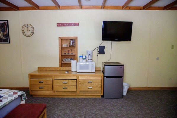 A room with a wooden dresser, a microwave, a refrigerator, a television, and a clock on the wall.
