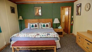 Une chambre à coucher avec un lit en bois, un banc, une table de chevet, une commode et une porte donnant sur une autre pièce.