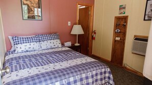 Quarto com colcha xadrez, mesa de cabeceira com abajur, porta com placa e prateleira na parede.