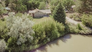 A house with a sloped roof, surrounded by dense greenery and a river. - C-105) Coyote Gulch Cabin (Sevier)