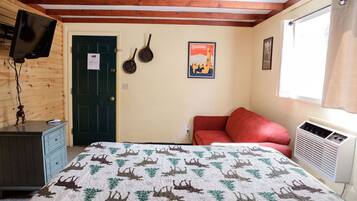 A room with a bed, a red sofa, a wooden dresser, a television mounted on the wall, a door, and a poster on the wall.
