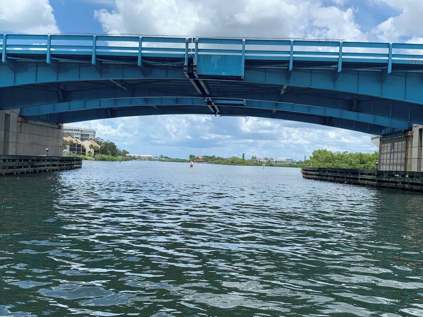 A blue bridge over a calm river with buildings on the opposite bank.