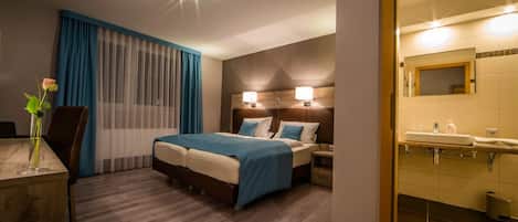 A hotel room with a bed, bedside tables, a desk with a vase, and a bathroom with a sink and mirror.