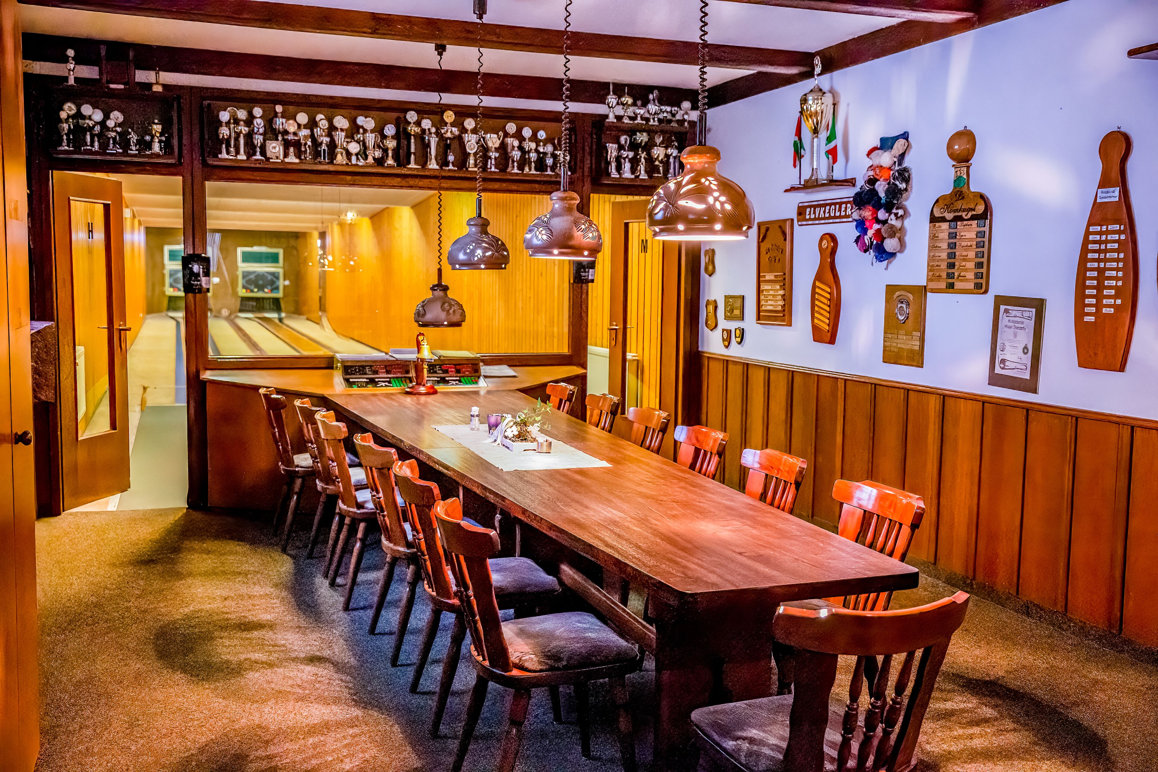 A dining area with a long wooden table, multiple chairs, hanging pendant lights, and a wall decorated with trophies and plaques.