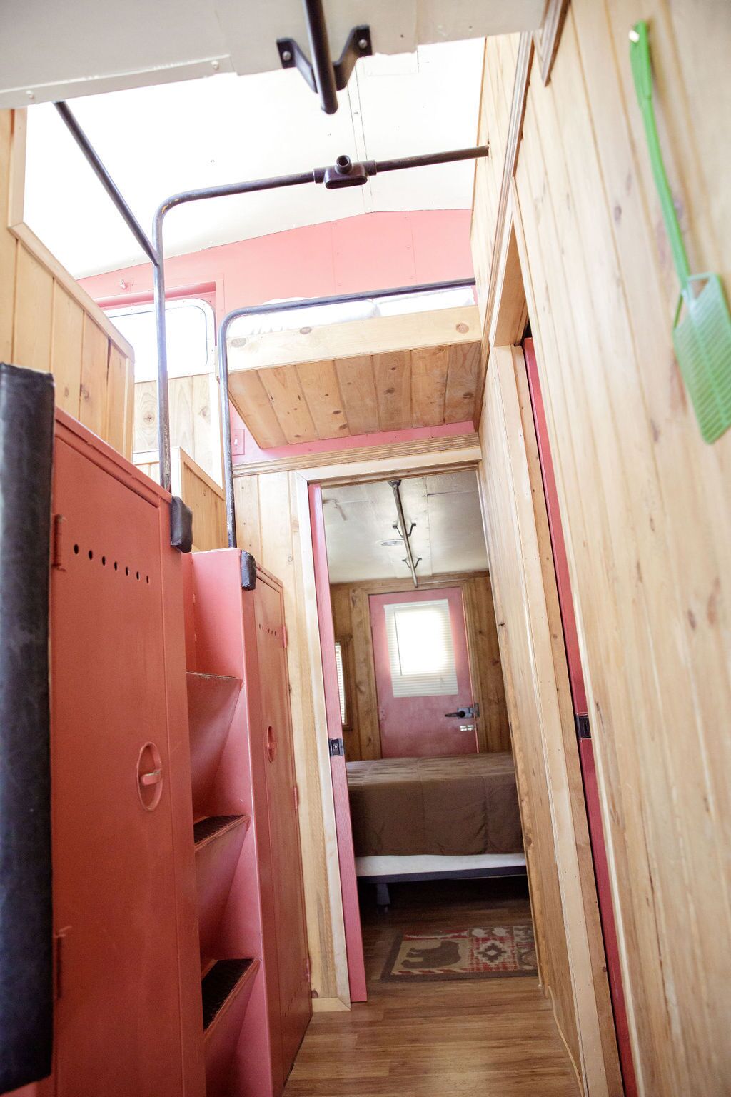Un pasillo estrecho con paredes y techo de madera. Se ve una cama a lo lejos, y un portalápices verde está en la pared derecha.