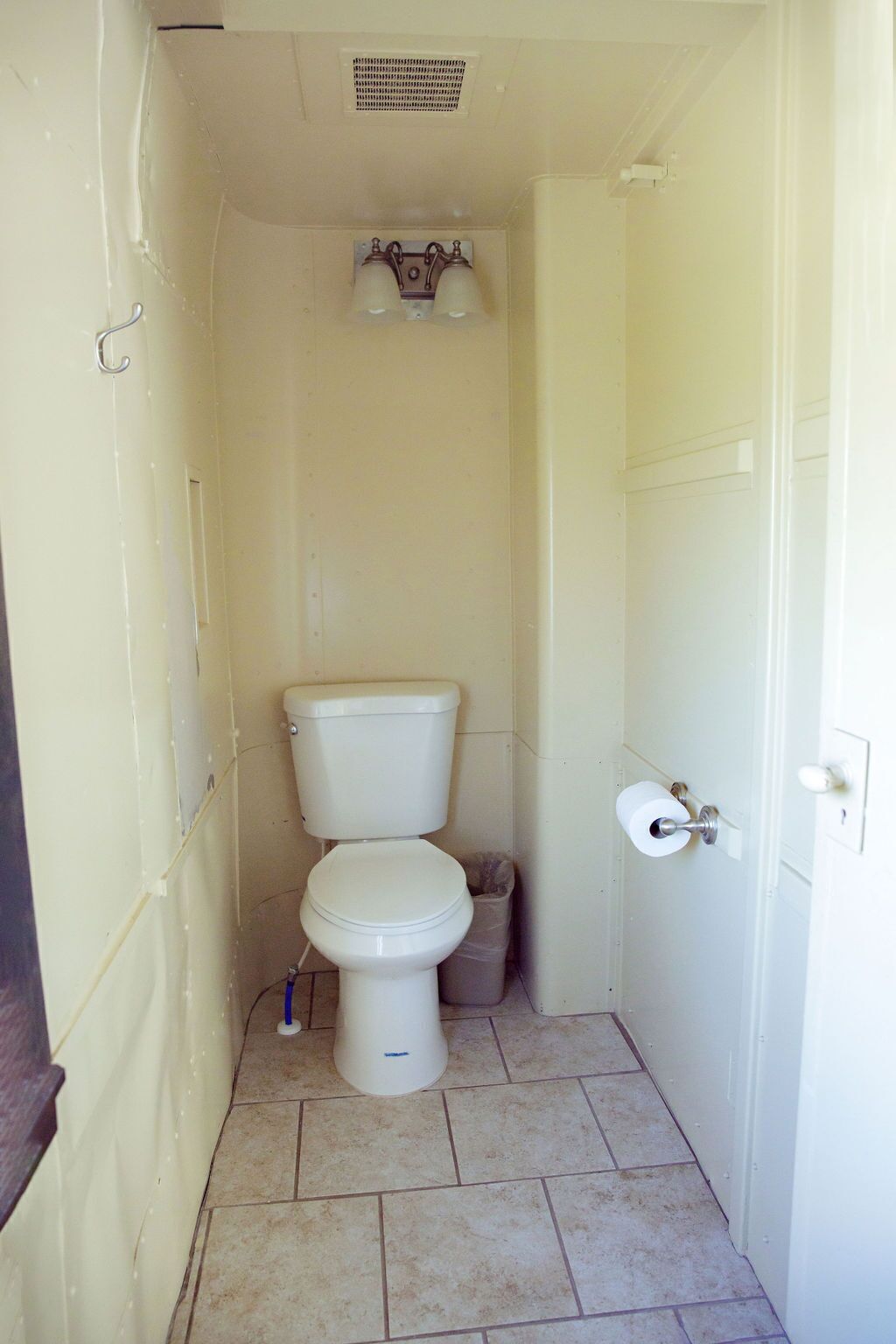 Une petite salle de bain étroite, équipée de toilettes, d’une poubelle et d’un luminaire fixé au mur.