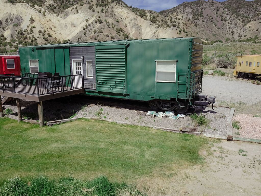 Un wagon de train vert, doté d’un petit porche et d’une fenêtre, situé dans une zone rurale avec des montagnes en arrière-plan.