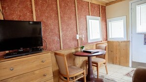Une pièce avec un téléviseur posé sur une commode en bois, une petite table avec des chaises et une fenêtre avec des stores.