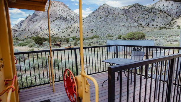 Une terrasse dotée d’une rambarde en métal, d’une table et de chaises. Au fond, on aperçoit des montagnes et une végétation clairsemée.