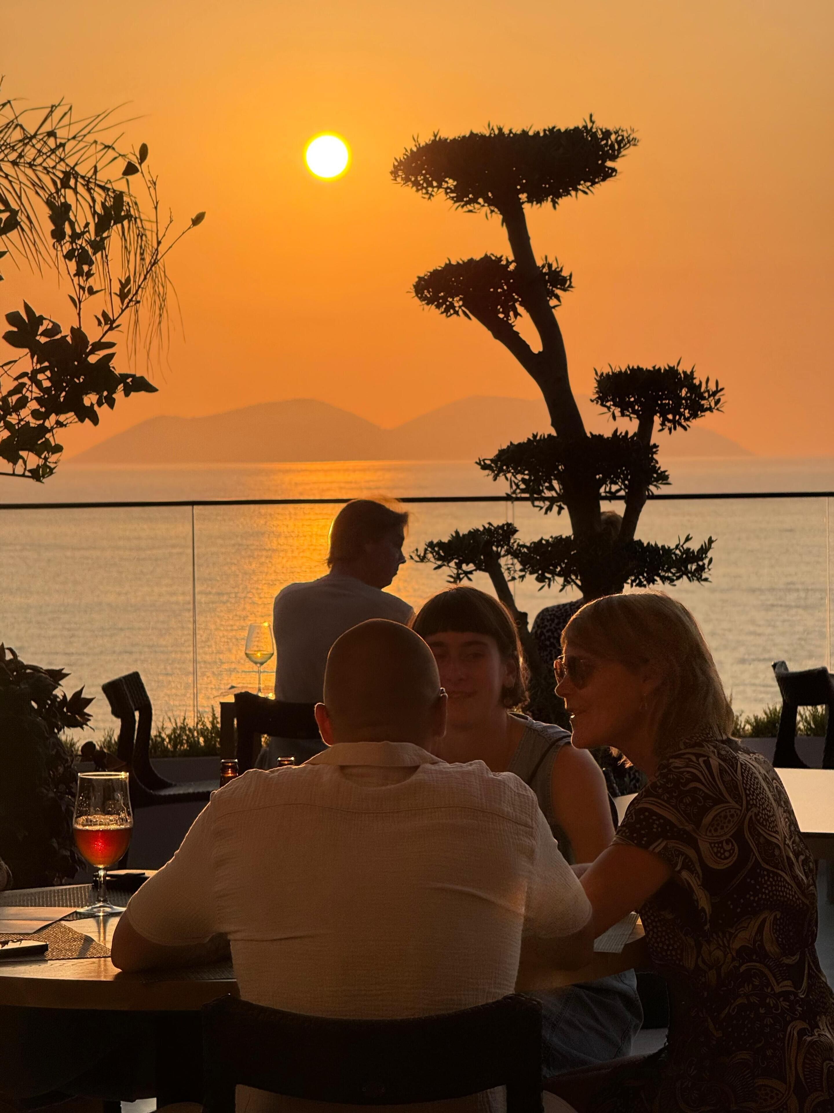 A sunset view from a terrace with a table and chairs, overlooking a body of water and distant mountains.