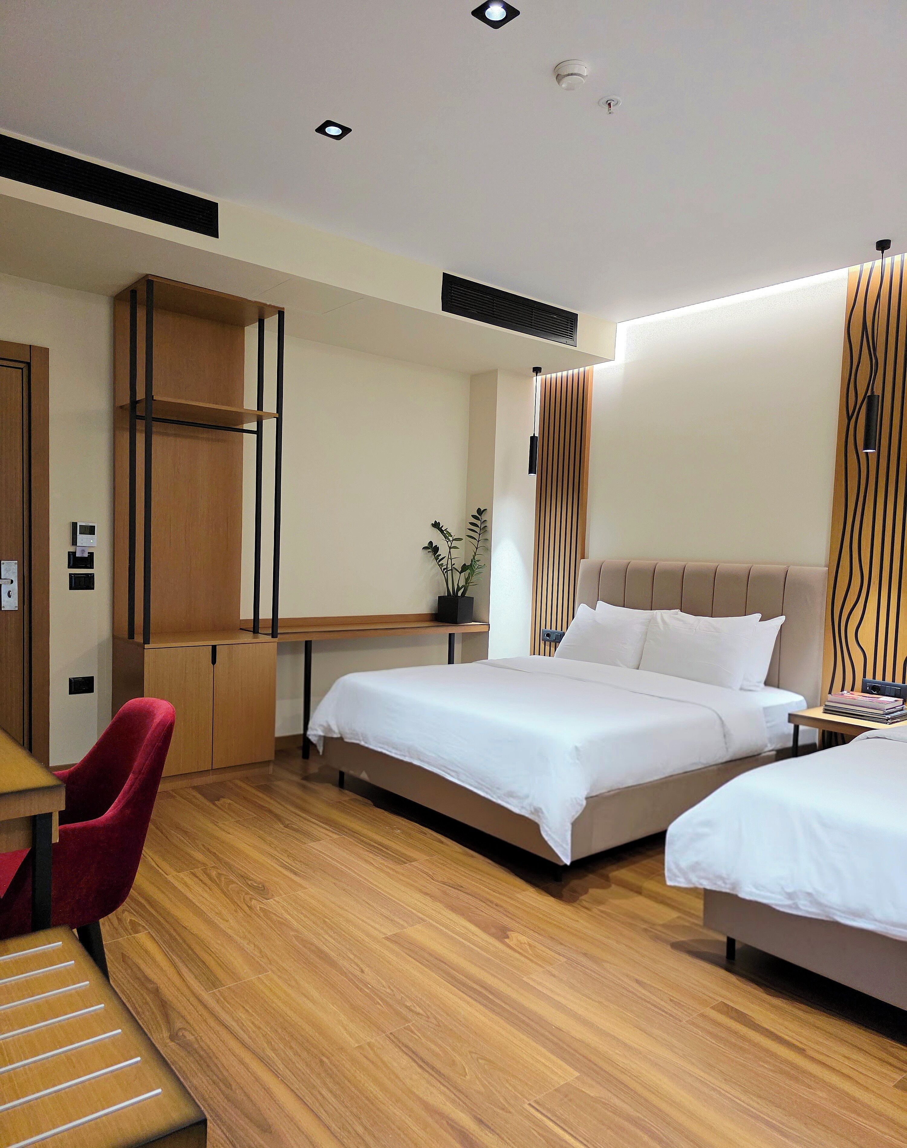 Une chambre d’hôtel moderne avec deux lits, un bureau, une chaise rouge et un plancher en bois.