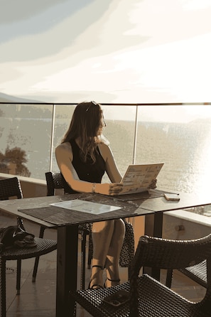 A woman sitting at a table with a menu, overlooking a body of water. - Valza Boutique Hotel (Vlorë)