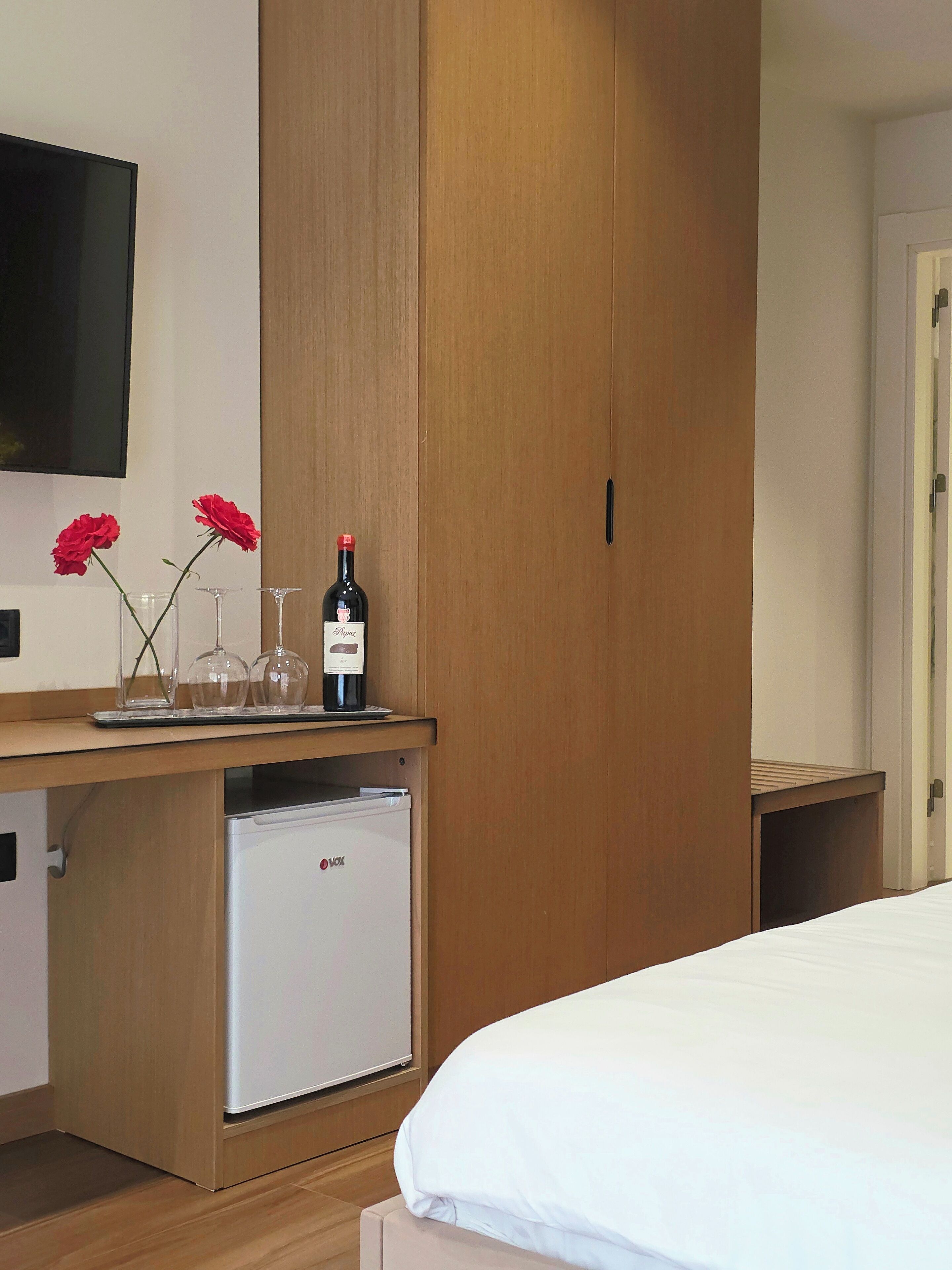 A hotel room with a bed, a wooden wardrobe, a small refrigerator, a TV mounted on the wall, and a wine bottle with glasses on the dresser.