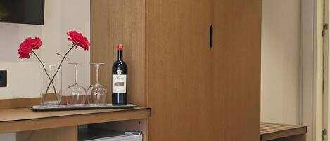 A hotel room with a bed, a wooden wardrobe, a small refrigerator, a TV mounted on the wall, and a wine bottle with glasses on the dresser.