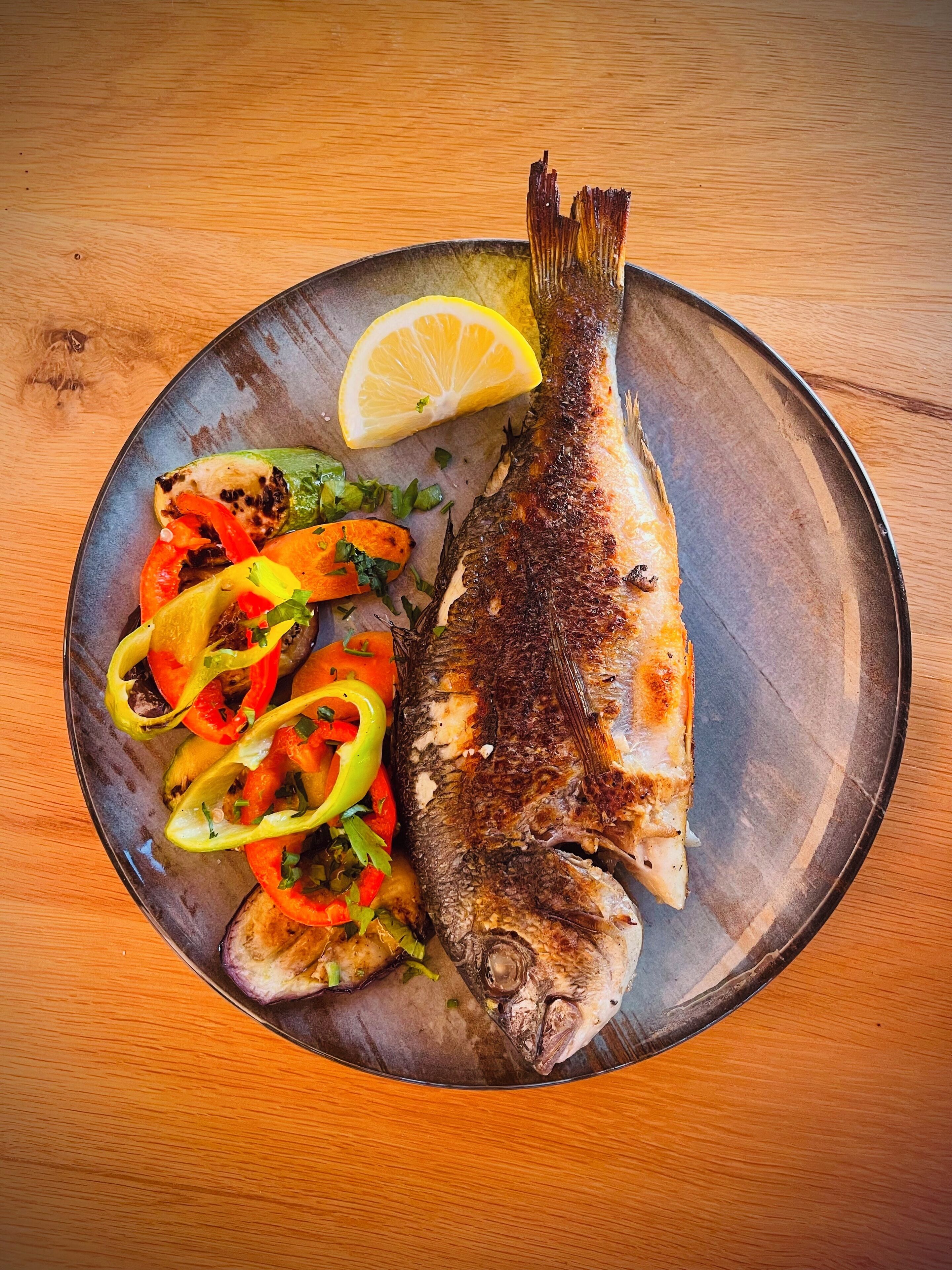 A whole grilled fish with lemon and a side of grilled vegetables on a plate.