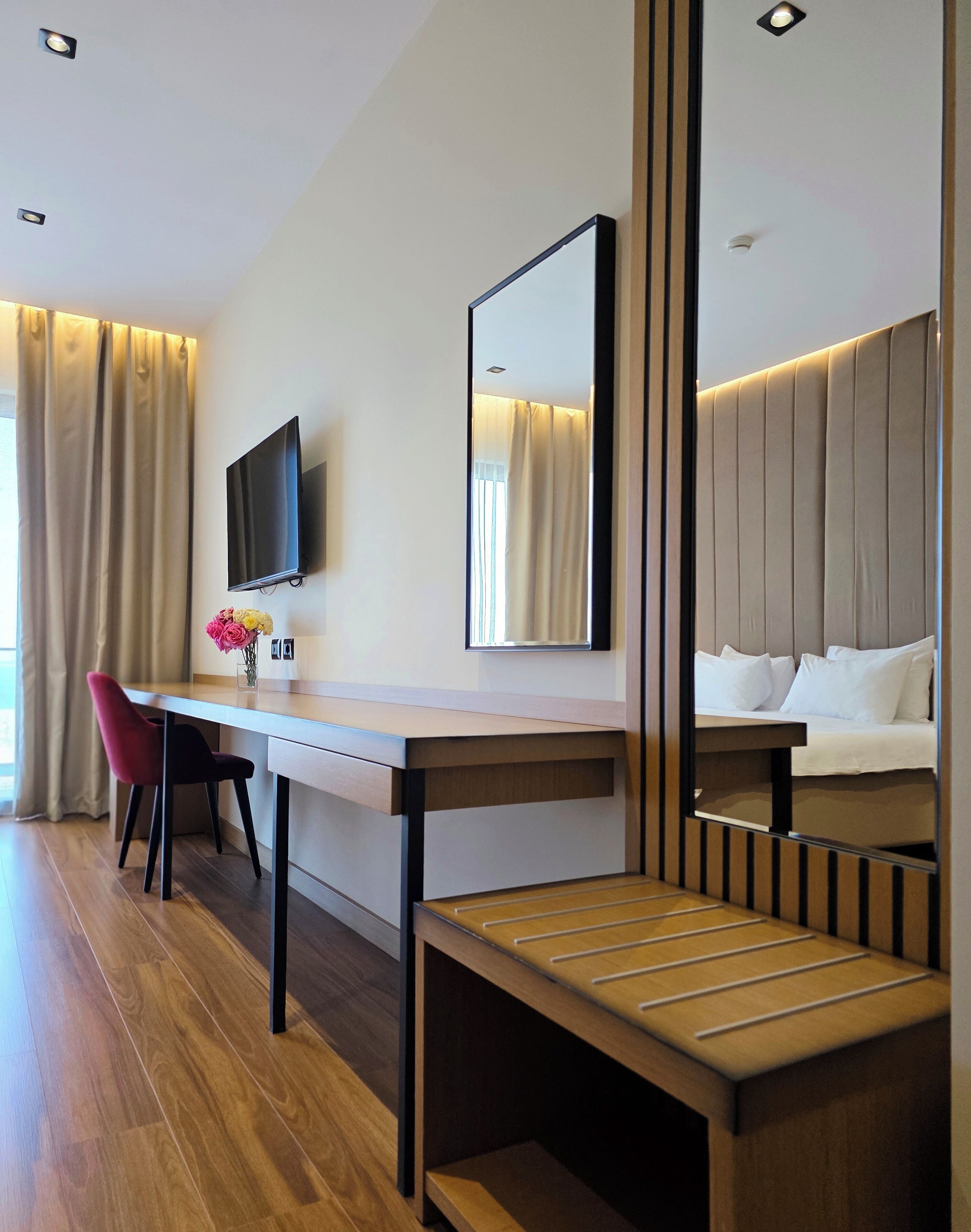 Habitación de hotel con un escritorio de madera, una silla, una cama con ropa de cama blanca, un televisor montado en la pared y un espejo que refleja parte de la habitación.