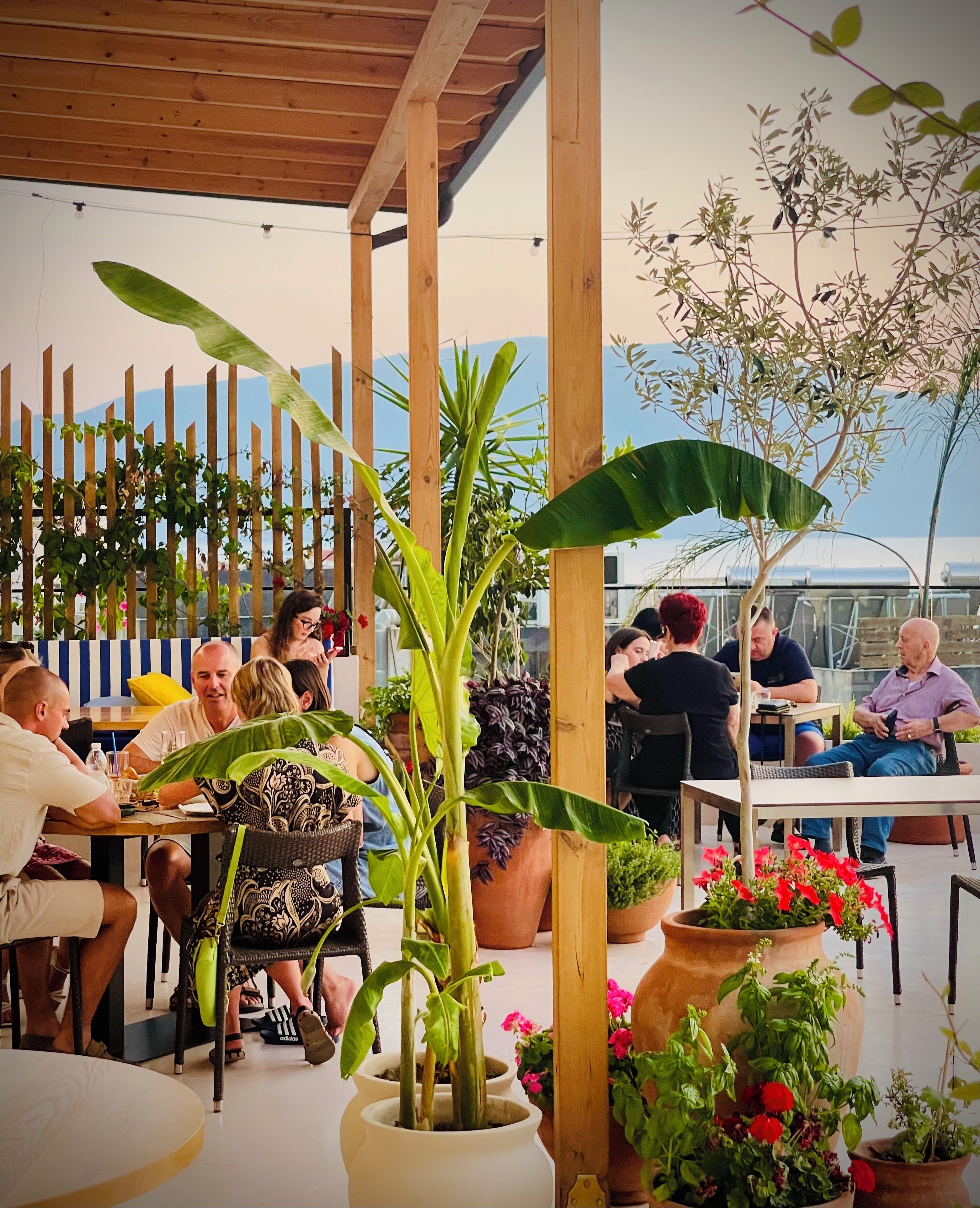 Een terras buiten met houten tafels en stoelen, omringd door plantenpotten en een houten hek.