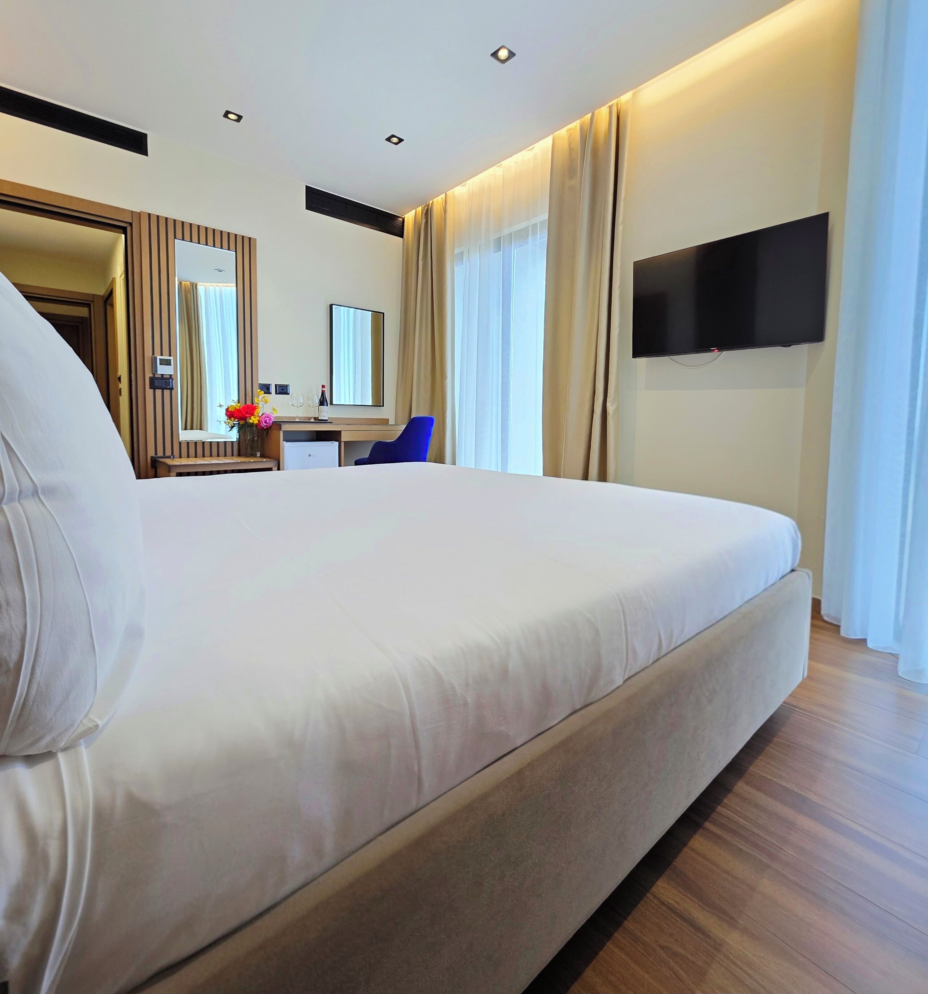 A modern hotel room with a large bed, a flat-screen TV mounted on the wall, a desk with a mirror, and a small refrigerator.