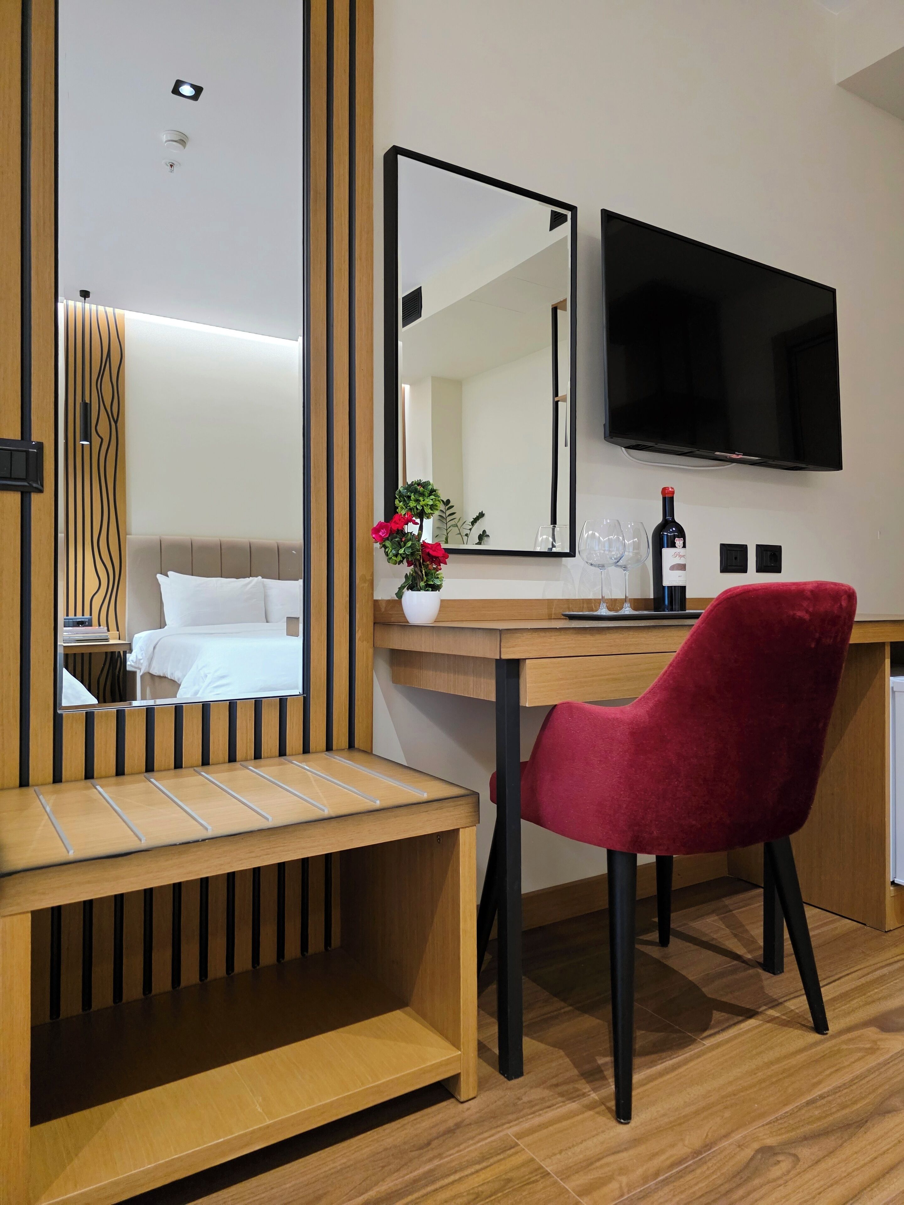 A hotel room with a wooden desk, a red chair, a large mirror, a flat-screen TV, a wine bottle, and a vase with flowers.