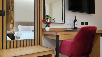 A hotel room with a wooden desk, a red chair, a large mirror, a flat-screen TV, a wine bottle, and a vase with flowers.
