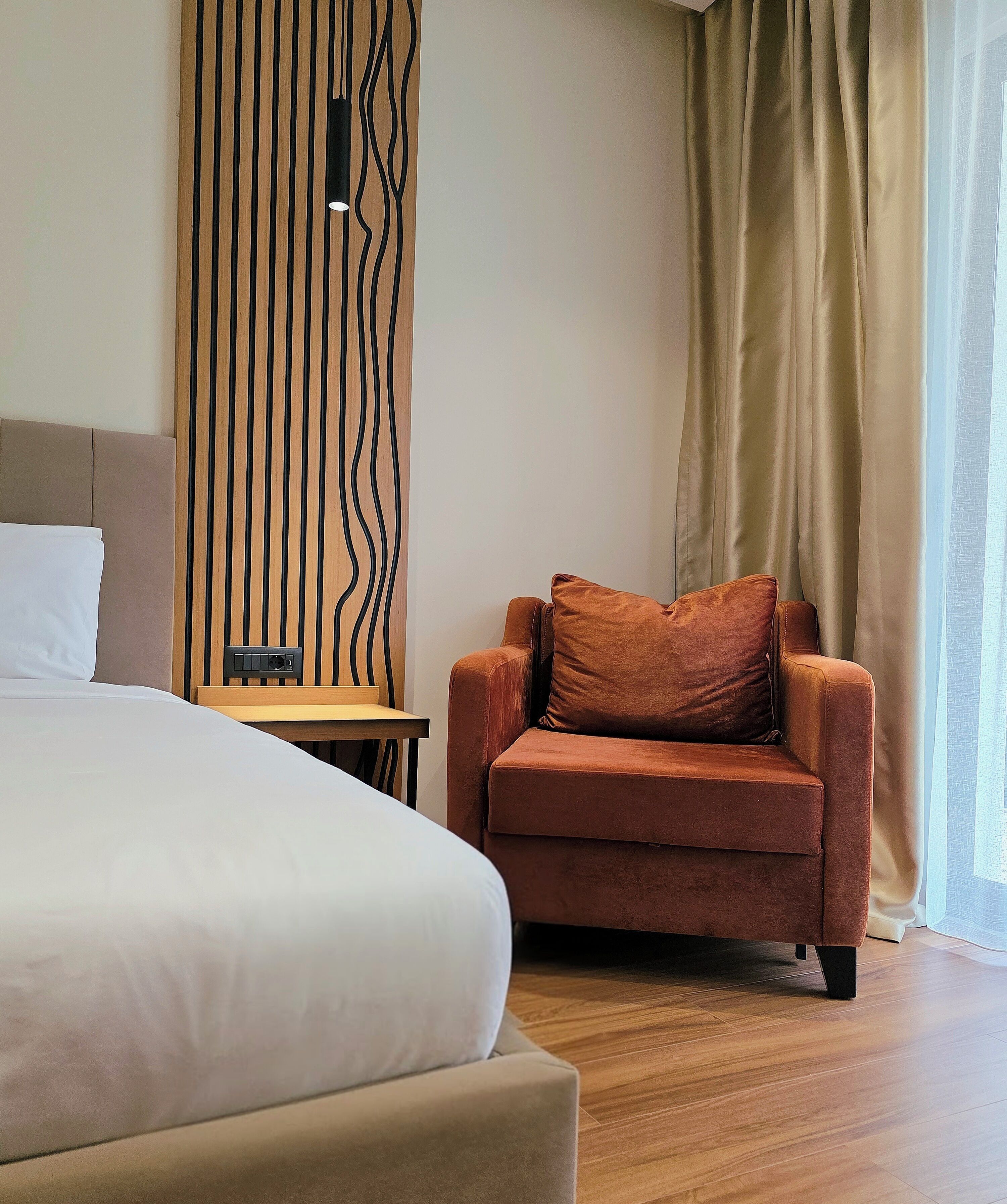 A hotel room with a large bed, a brown armchair with a pillow, a wooden side table, and a wall with a vertical wooden panel design.