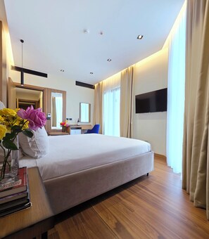 A modern hotel room with a large bed, wooden flooring, a desk with a chair, a TV mounted on the wall, and a window with curtains. - Valza Boutique Hotel (Vlorë)