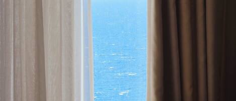 A room with a bed and a view of the sea through sheer curtains.
