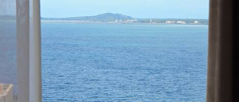 Vista al mar desde una ventana, con una masa de tierra distante y vegetación en la parte baja.