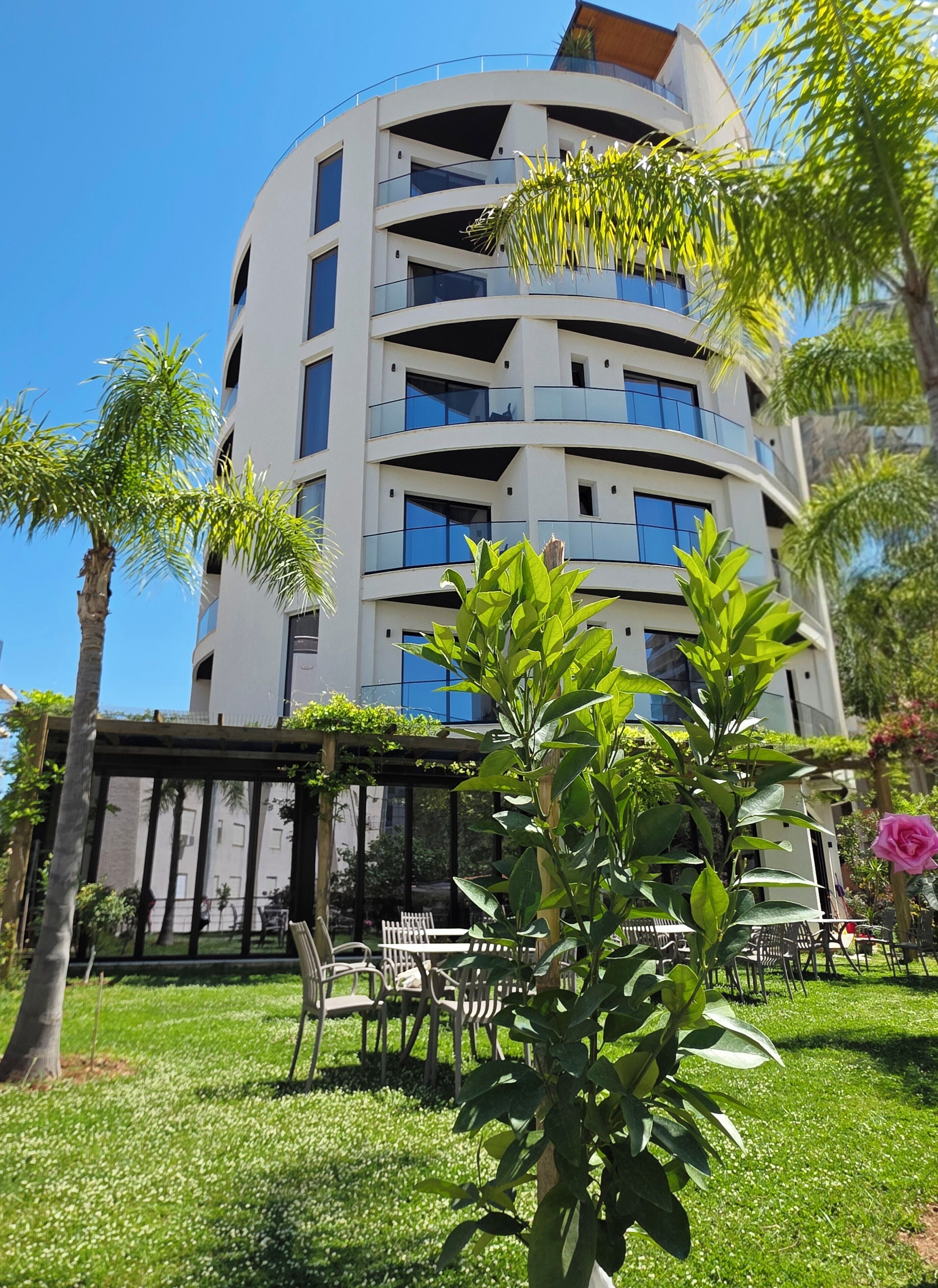Un edificio de varios pisos con balcones, rodeado de vegetación y mobiliario exterior.