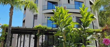 Un edificio de varios pisos con balcones, rodeado de vegetación y mobiliario exterior.